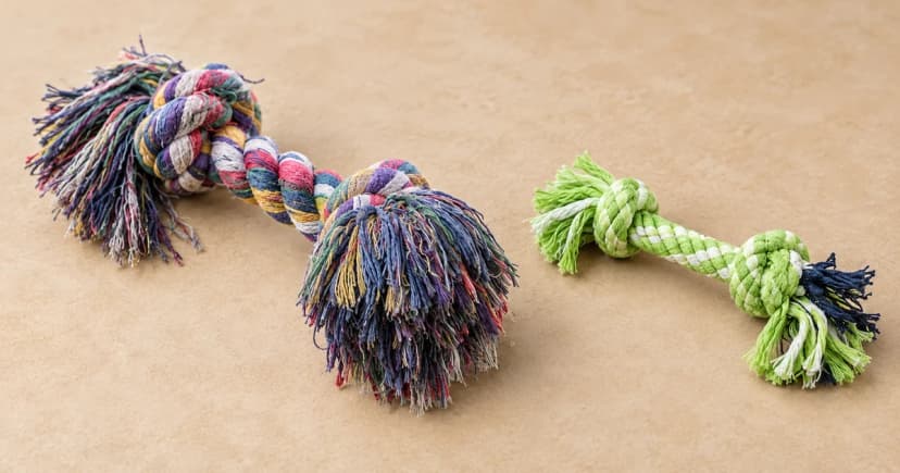 Two separate knotted rope toys, one multicolored and one green and navy, on a sand-colored tabletop.