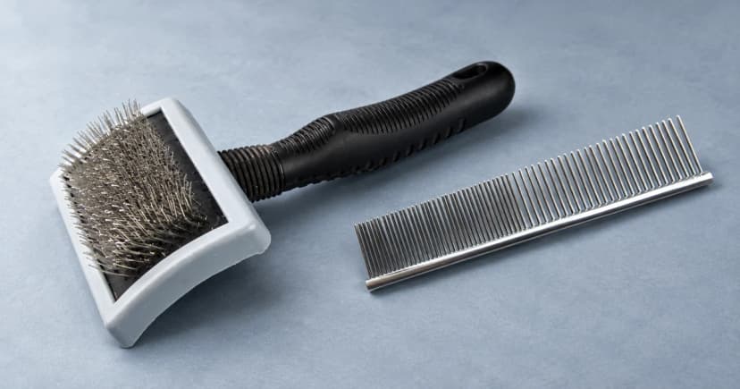 A wire slicker brush with a black handle in front of a separate metal comb on a blue-gray tabletop.