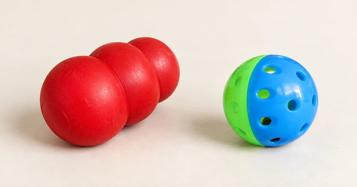 Blue-green perforated ball beside a red rounded toy lying on a pale tabletop.