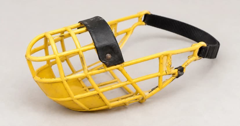 A yellow open-basket muzzle with a dark nose bridge and loose rear band rests on a pale tabletop.